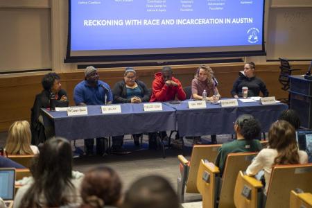 A group of people sit behind a table on a panel in front of a classroom of students